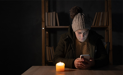 Kvinna med mössa och jacka sitter i mörkt rum och ser på telefonen, ett ljus på bordet