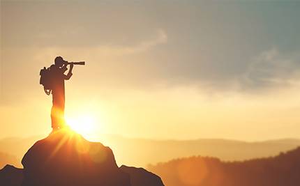 Man med kikare på en fjälltopp mot himmel med starkt solljus